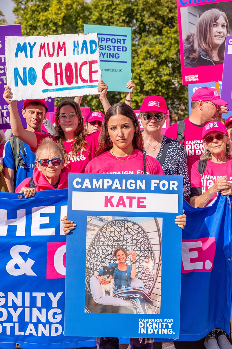 Portrait event photograph of Dignity in Dying supporters holding campaign placards and a large blue campaign board during a Westminster public affairs event