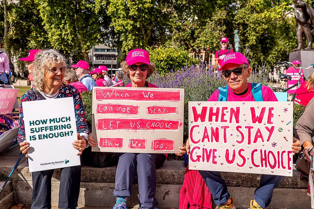 Dignity in Dying campaign supporters seated with handmade placards during a Westminster event, showing personal messages and public affairs campaign atmosphere